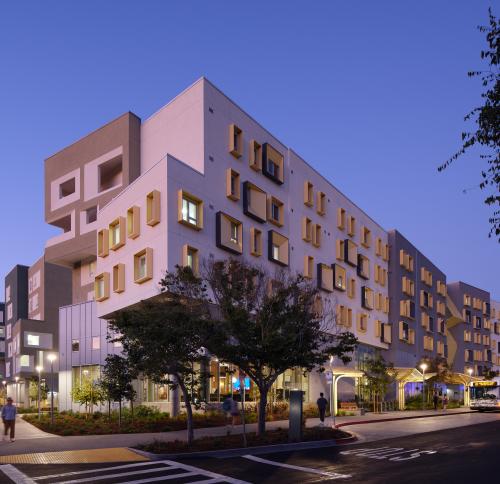 Exterior view of a multi-story student housing complex at dusk, featuring gray and white façades, projecting gold window surrounds, landscaped sidewalks, and a bus stop along the street.