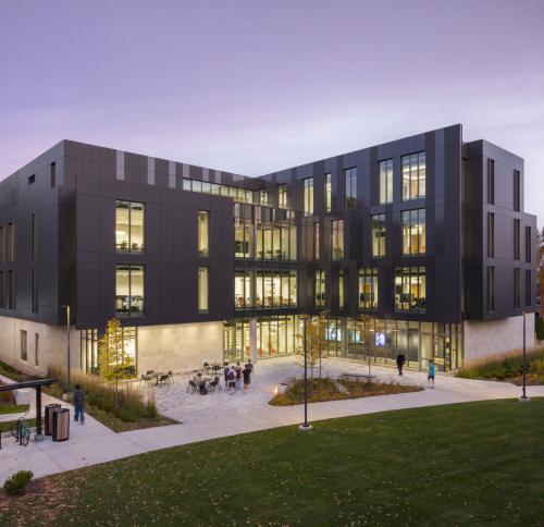 Exterior view of the academic building overlooking a landscaped courtyard, with transparent façades revealing interior makerspaces and collaborative learning areas as students move through the campus.