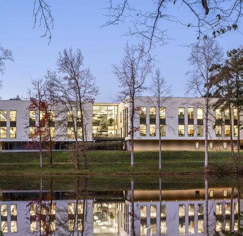 A contemporary white building situated on a grassy bank, surrounded by bare trees and reflected in a still lake.
