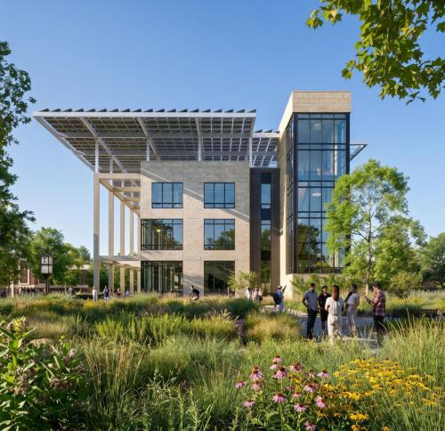 A contemporary academic building with large glass windows and a prominent solar array, surrounded by green spaces and mature trees.