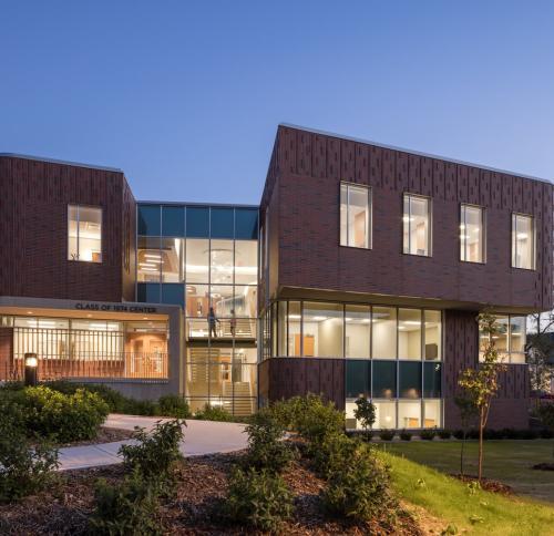 A modern, multi-story brick building with large glass windows, illuminated from within at dusk, featuring the "Class of 1974 Center."