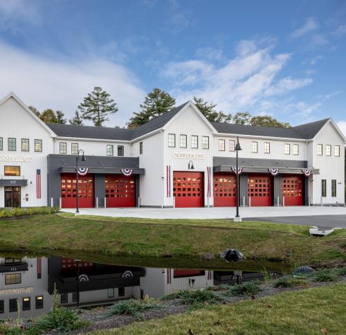 Exterior view of the station. A long white two story building with pitched New England style roof and features 6 large red garage doors