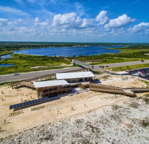 A coastal facility featuring two connected wooden structures with flat, white roofs. The building includes a covered porch area, large windows, and is flanked by a ground-mounted solar array on one side and extensive wooden boardwalk ramps on the other.