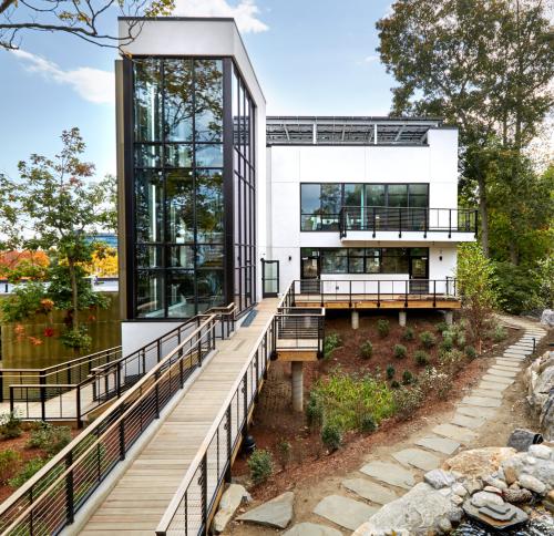 An exterior view of a contemporary, multi-level white building surrounded by trees, with a prominent glass stairwell and a wooden ramped walkway.