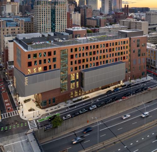 An aerial photo of the school. A seven story building with a rooftop green space, two silver volumes of building sticking our to the left and middle, and a two tone brick facade featuring windows with irregular protruding frame with bold colors of red, yellow, green, and blue. The school occupies a narrow strip of a city block that sits adjacent to the visible subterranean highway.