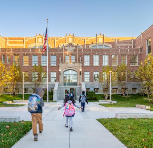 A historic brick school building with large windows and an ornate central entrance. A flagpole stands in front of a landscaped courtyard with walkways leading to the doors. Students with backpacks are walking toward the entrance.