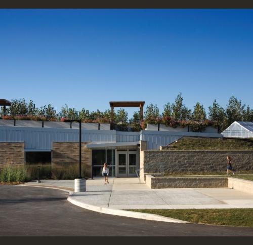 A low-profile, modern institutional building with a flat roof supporting a green roof, a central glass entrance, and an adjacent greenhouse structure.