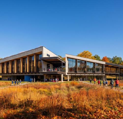 An architectural view of a long, modern structure featuring a mix of wood and glass, surrounded by a natural landscape with fall foliage.