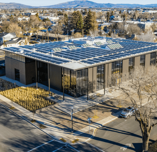 A aerial view of a modern, flat-roofed building with a large solar panel array covering the roof, set against a backdrop of distant mountains.