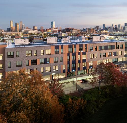 Four‑story Roxbury Prep building at dusk with the Boston skyline in the background and fall trees in the foreground.