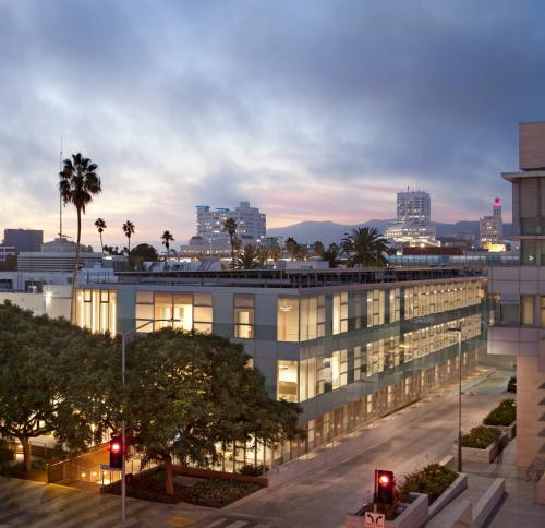 A contemporary multi-story building with large windows, glowing from within against a twilight sky. Palm trees and streetlights are visible in the foreground, with a distant city skyline and mountains behind.