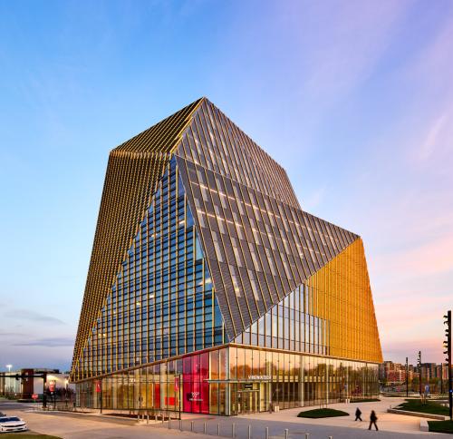 A low-angle view of a modern, geometric building, featuring a glass facade with gold-colored accents against a twilight sky.